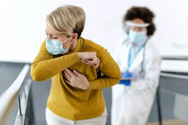 Female patient coughing into elbow while wearing protective face mask at he hospital. Doctor is standing in the background.