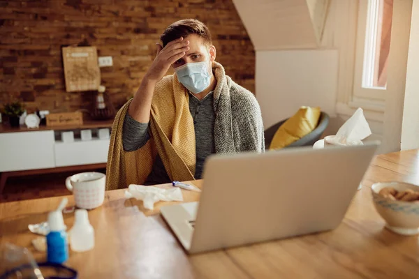 Sick man having fever and talking with a doctor online over laptop while wearing protective face mask at home. 