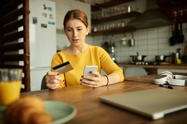 Young woman using credit card while checking her online bank account over mobile phone at home. 