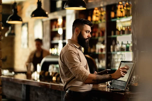 Young bartender using cash register at bar counter in a pub.