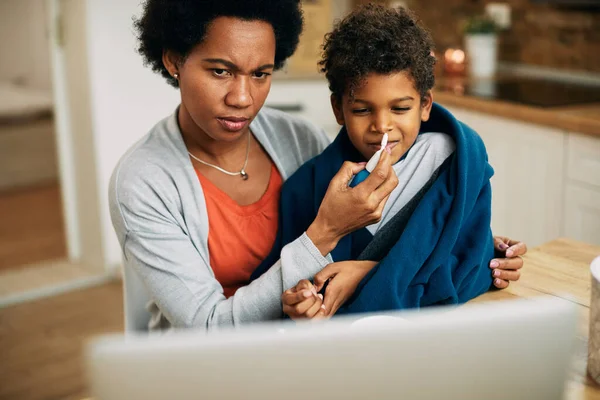 Black mother applying nasal spray to her sick son while following instructions on a computer at home.