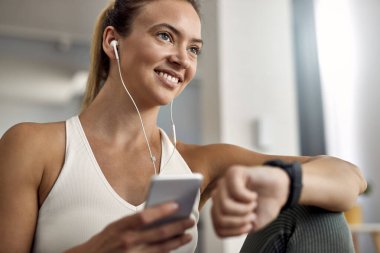Young happy sportswoman using smart phone while checking the time on her wristwatch at home. 