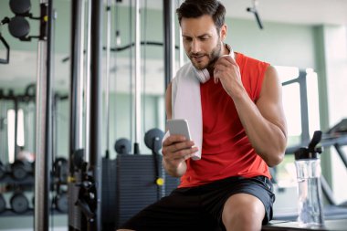 Athletic man taking a break from exercising and texting on mobile phone in a gym. 