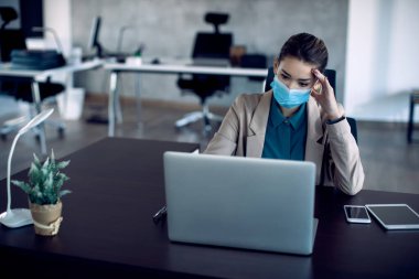 Young businesswoman feeling worried while reading bad news on a computer in the office. She is wearing face mask due to COVID-19 pandemic.