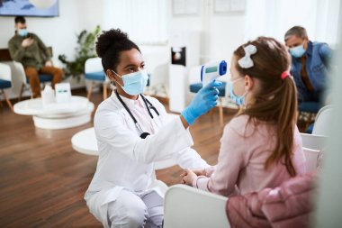 Black doctor using infrared thermometer and measuring temperature of her small patient at dental waiting room dur to COVID019 pandemic.