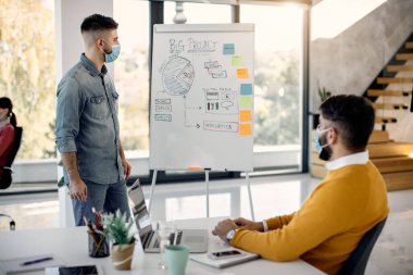 Male entrepreneur wearing face mask while presenting business strategy on whiteboard to a colleague in the office.