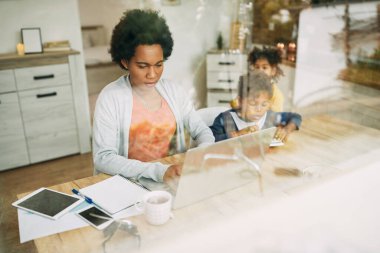 Black female entrepreneur working on a computer while being with her children at home. The view is through the glass. Copy space.