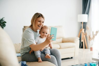 Young mother using smart phone and taking selfie with her baby son at home.