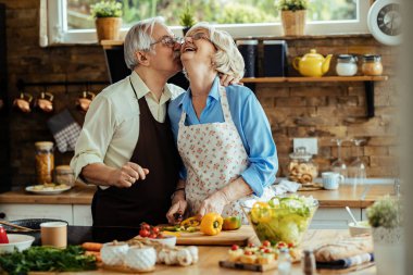 Happy senior couple having fun while cooking together. Man is kissing his wife. 