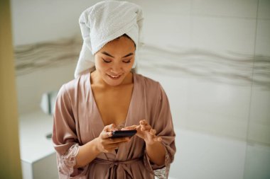 Bornozlu gülümseyen Asyalı kadın banyoda akıllı telefon kullanıyor..