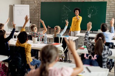 Happy African American teacher holding a class and aiming at her students who are raising their hands to answer the question in the classroom. 