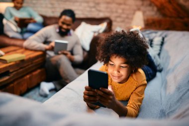 Smiling black girl using mobile phone while relaxing on the sofa at home. Her parents are in the background.