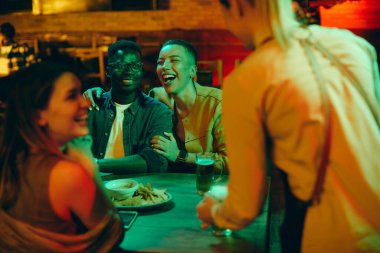 Happy African American woman and her friends laughing and having fun while waitress is serving them drinks during their night out in a bar. 