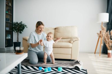 Young happy mother assisting her baby son with his first steps at home. Copy space.