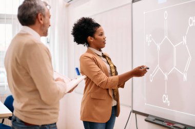Black science professor giving chemistry lecture to her students while standing in front on whiteboard at lecture hall.