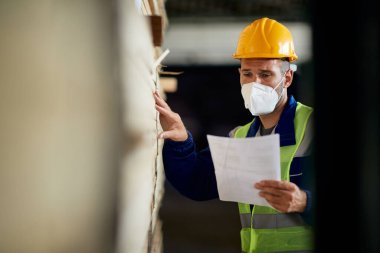 Warehouse worker with face mask going through inventory list while checking stock of timber at storage compartment. Copy space.