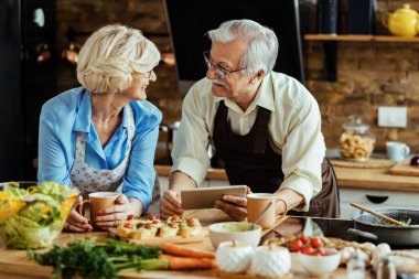 Happy mature couple using digital tablet and talking while preparing lunch in the kitchen.