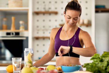 Happy athletic woman checking time on her wristwatch while preparing fruit smoothie in the kitchen. 