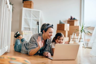 Happy single mother and daughter using laptop and greeting someone during video call while relaxing on the floor at their new home. 