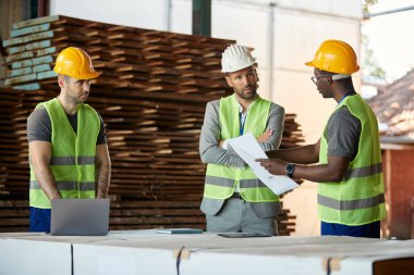 Warehouse workers and their manager talking while analyzing paperwork at lumber department.