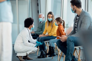 Black female pediatrician communicating with little girl and her mother in a waiting room at medical clinic. All of them are wearing face masks due to coronavirus pandemic. 