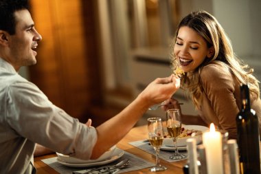 Happy woman having fun while being fed by her boyfriend during a meal in dining room.