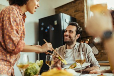 Young happy man sitting at dining table while his African American girlfriend is serving lunch. 