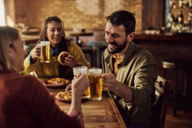 Group of happy friends toasting with beer while having lunch in a pub. Focus is on man. 