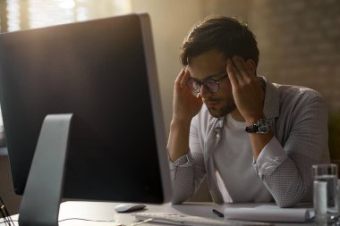 Young businessman man holding his head in pain and feeling exhausted after working on desktop PC in the office.