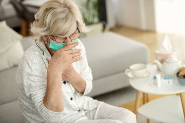 Mature woman with protective face mask coughing while feeling chest pain at home during COVID-19 epidemic. 