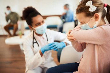 Little girl wearing face mask and coughing into elbow while talking to a doctor in waiting room. 
