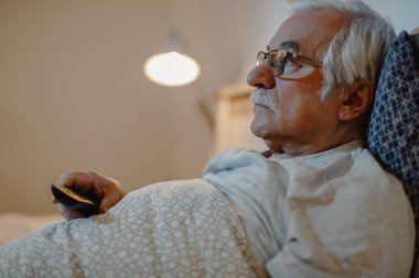 Mature man lying down in bed and watching TV at night. 