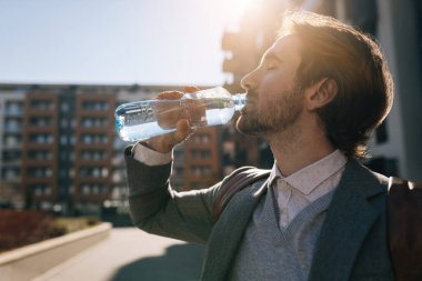 Side view of young businessman refreshing himself and drinking water in the city. 