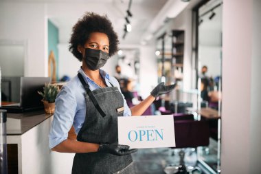 African American hairdresser wearing face mask while holding open sign and welcoming her customers at hair salon.