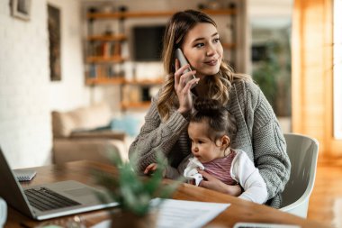Smiling mother holding small daughter in her lap while talking on the phone at home. 