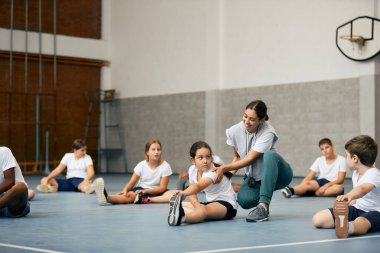 Group of school children exercising during physical activity class at school gym. Happy teacher is assisting girl with stretching exercise. 