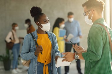 Black female student ad her friends wearing protective face masks while communicating at university hallway. 