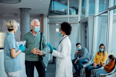mature man and healthcare workers communicating in a hallway at medical clinic while wearing protective face masks. 