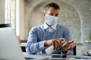 Male entrepreneur wearing protective face mask while using hand sanitizer in the office. 