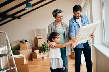 Happy parents and their daughter looking at wrapped painting while moving into their new home. 