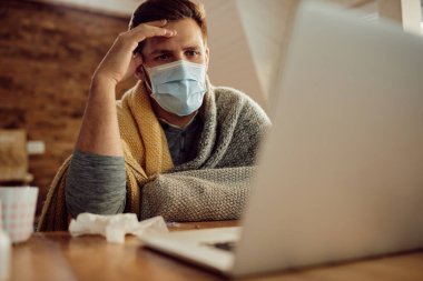 Distraught man using computer while wearing face mask and sitting wrapped in a blanket at home.