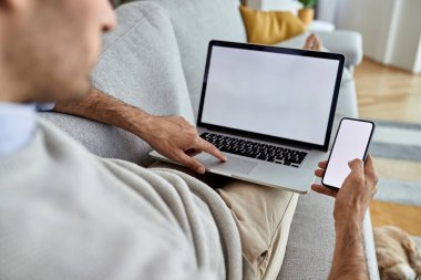 Close-up of freelance worker using mobile phone and laptop while relaxing on the sofa at home. Copy space. 