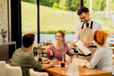 Young happy woman and her friends communicating while making order from a waiter in a cafe. 