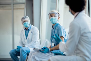 Group of medical experts wearing face shields and masks while communicating in a hallway and sitting apart from each other due to coronavirus pandemic. Focus is on nurse. 