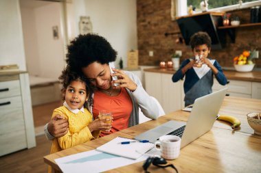Affectionate black mother embracing her small daughter while communicating on cell phone and working at home. 