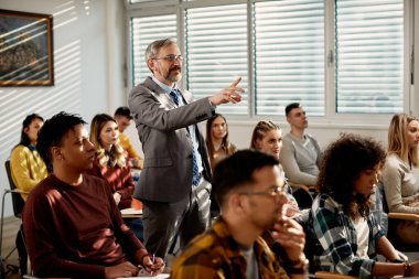 Mature professor pointing at something while holding lecture to his students in the classroom. 