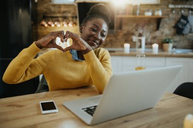Happy African American woman using computer while dating online and showing heart shape to someone.