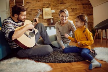 Mid adult father playing acoustic guitar while relaxing with his family at home. Focus is in mother. 