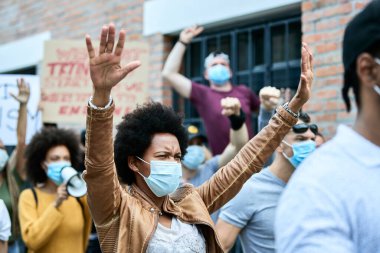 Displeased black woman wearing protective face mask while protesting with crowd of people on city streets during COVID-19 epidemic. 