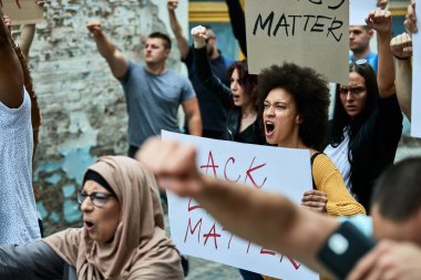 Large group of displeased people shouting on Black Lives Matter protest. Focus is on young black woman carrying a banner. 